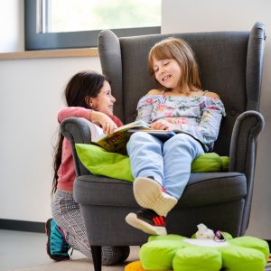 Zwei M&auml;dchen lachen und lesen ein Buch. Ein M&auml;dchen sitzt auf einem Sessel, das andere M&auml;dchen kniet neben dem Sessel.